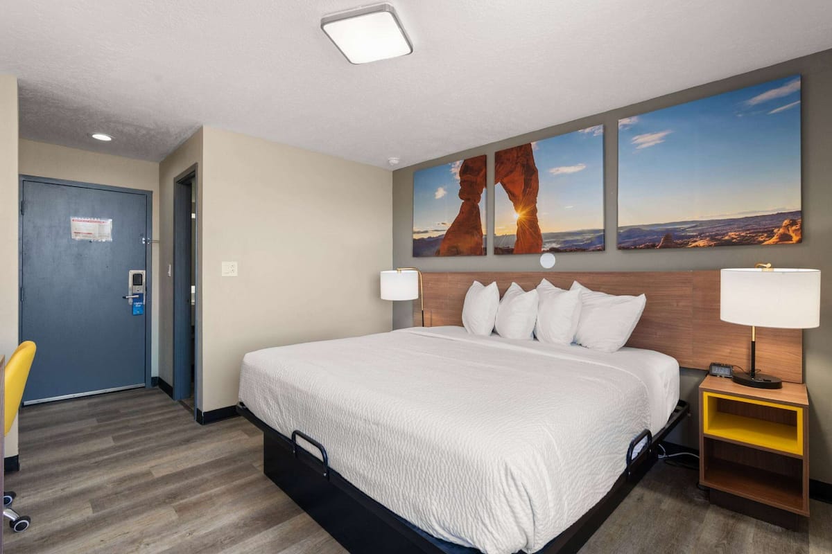 A hotel room with a large bed, a desk with a lamp, a chair, and a door.