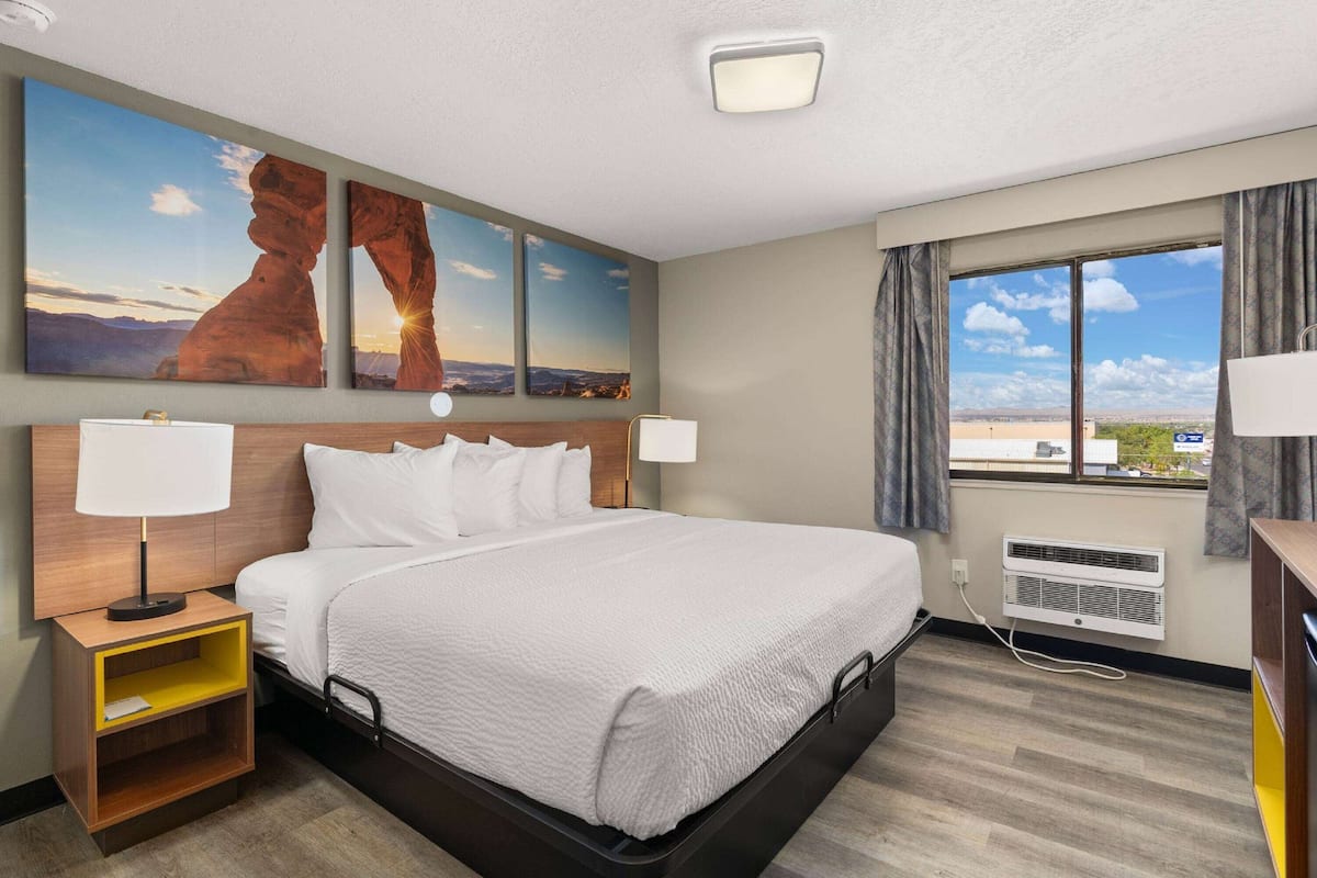 A hotel room with a large bed, two wall-mounted lamps, a nightstand, a window with curtains, and a wall-mounted air conditioner.