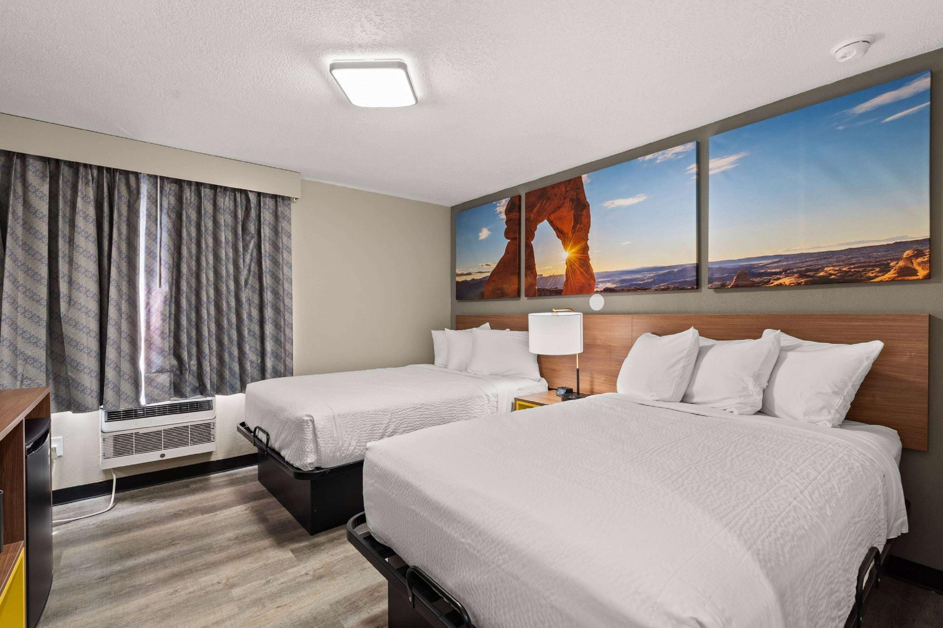 A hotel room with two beds, a large window with curtains, a wall-mounted air conditioner, and a painting of a desert landscape.