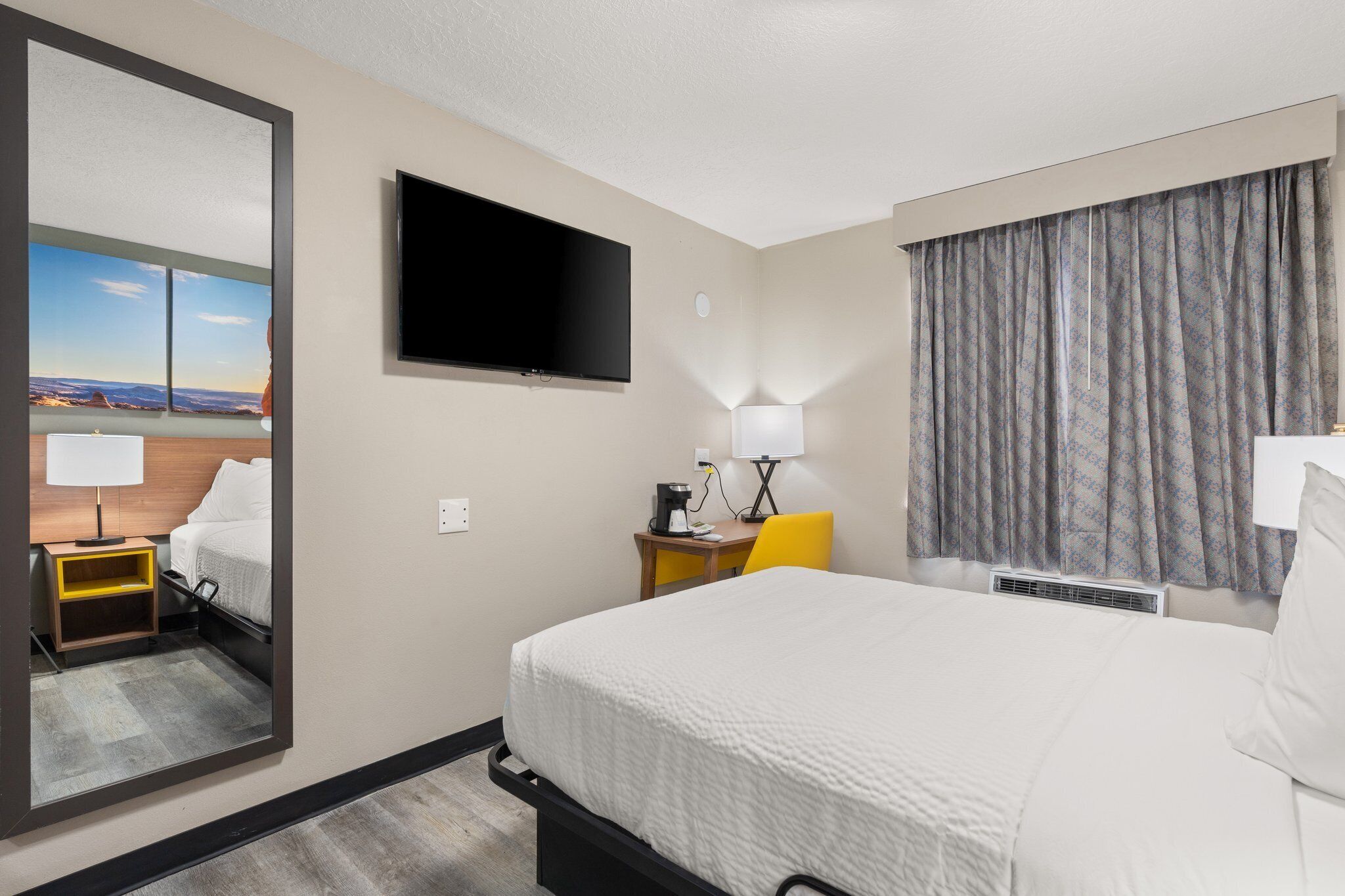 A hotel room with a bed, a desk with a lamp, a flat-screen TV mounted on the wall, and a large window with a view of a landscape.