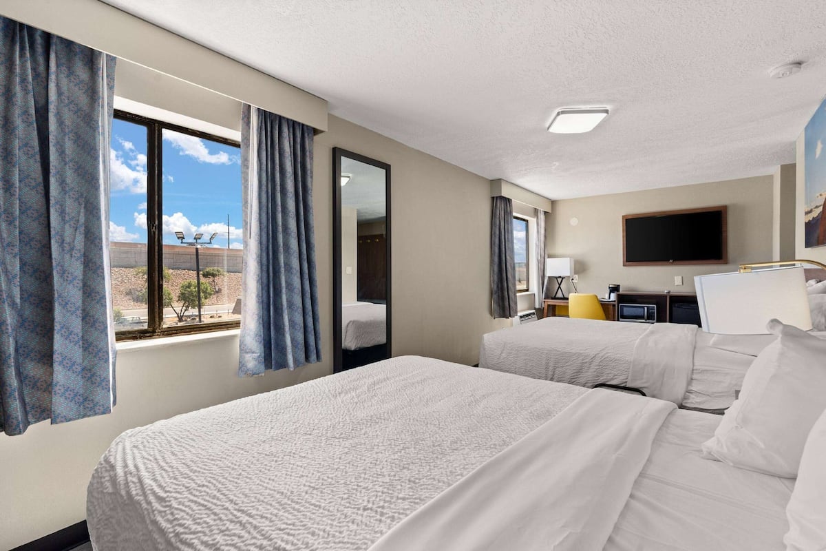 A hotel room with two beds, a flat-screen TV, a desk with a computer, and a window with curtains.