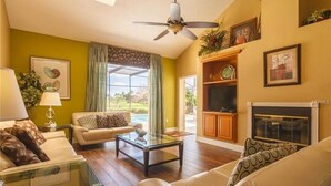 Ein Wohnzimmer mit einer Couch, einem Couchtisch und einem Kamin. Es gibt einen Deckenventilator, einen Fernseher und ein Fenster mit Vorhängen.