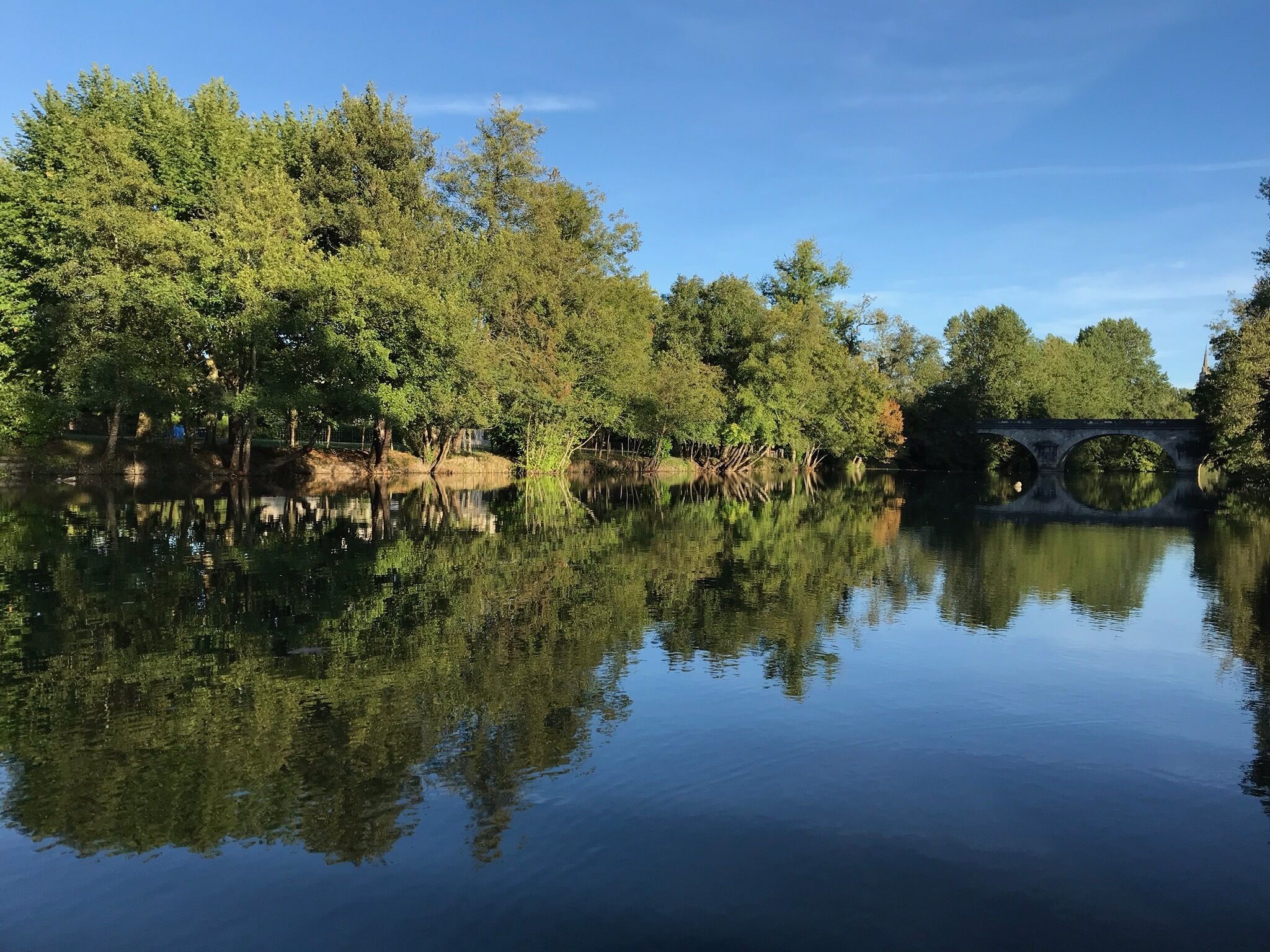 A calm river with a stone bridge and dense trees reflecting in the water.