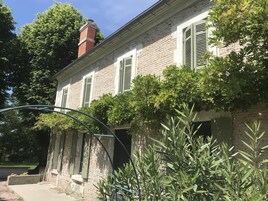 Un edificio de ladrillo de dos pisos, con contraventanas verdes, una chimenea visible y un jardín con diversas plantas.