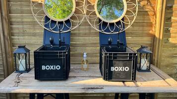Una mesa de madera con dos cajas negras etiquetadas "BOXIO" y dos faroles negros, frente a una valla de madera con espejos redondos en la parte superior.