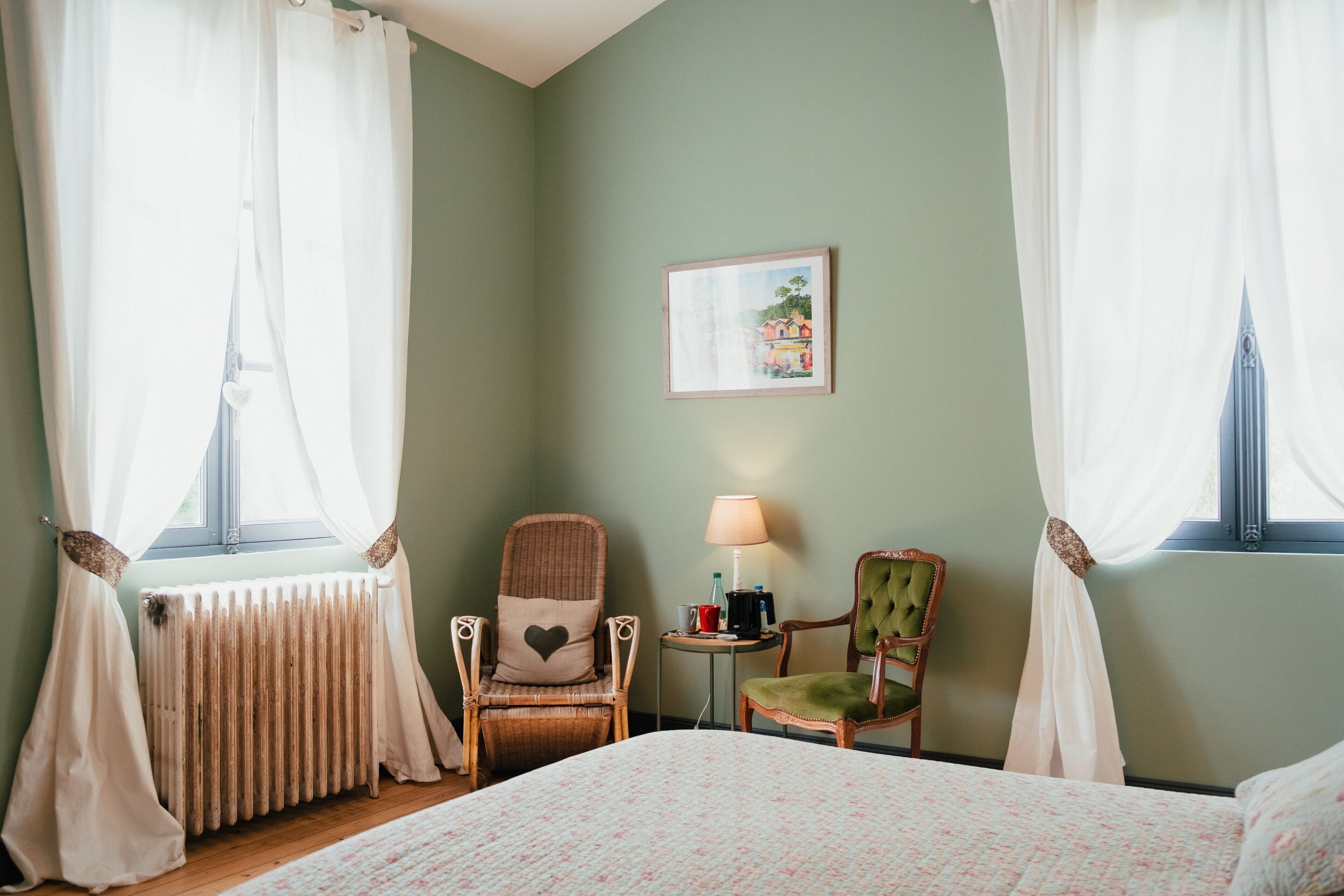 Quarto de hotel com cama, duas cadeiras, mesa com abajur, quadro na parede e janela com cortinas.