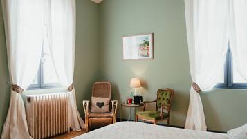 Quarto de hotel com cama, duas cadeiras, mesa com abajur, quadro na parede e janela com cortinas.