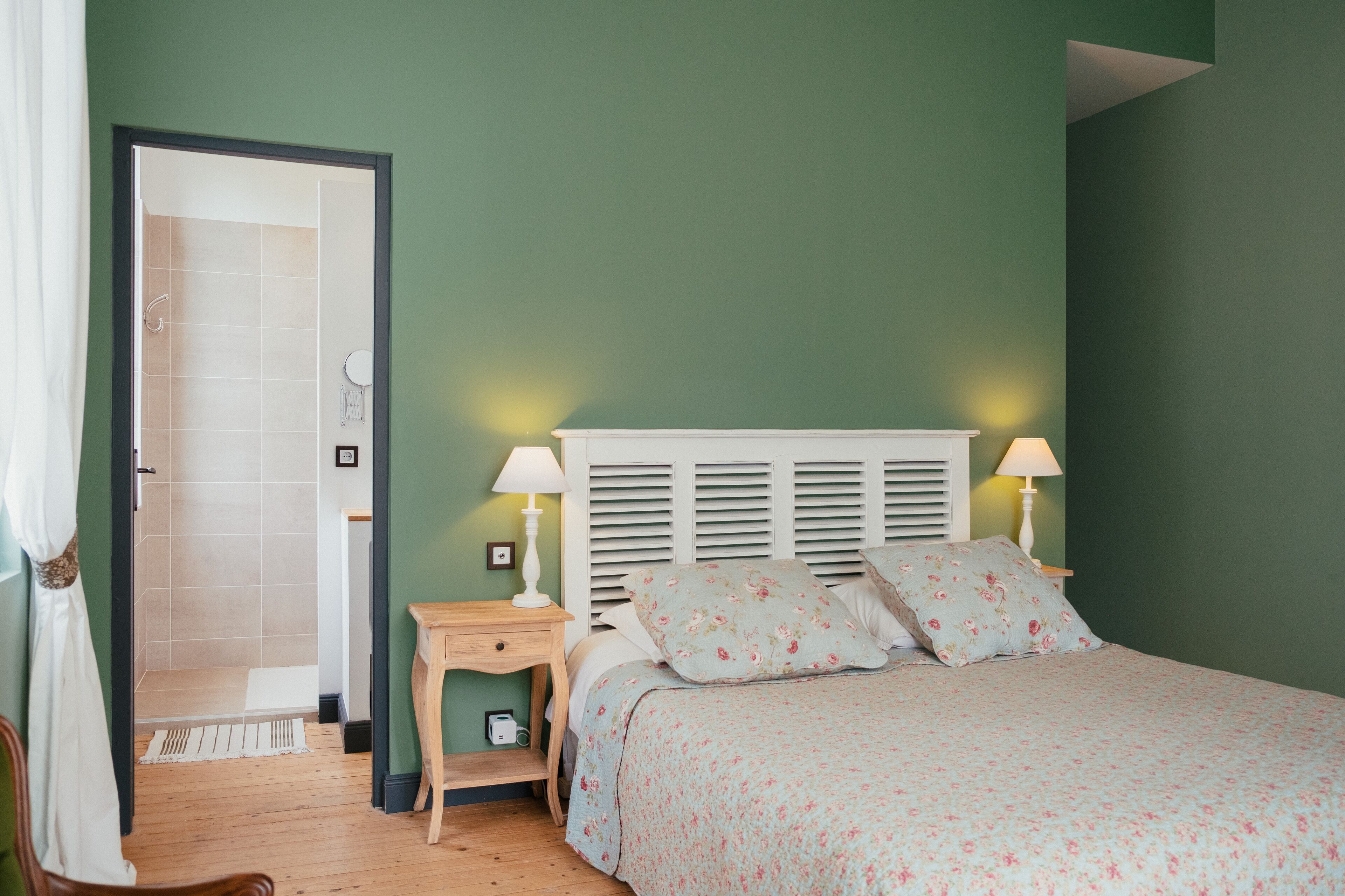 A bedroom with a bed, two bedside tables, a nightstand, a window with blinds, and a door leading to a bathroom.