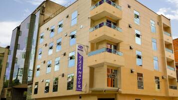 Un hotel de varios pisos con fachada plana, balcones en cada nivel y un llamativo cartel púrpura que indica el nombre y la dirección.