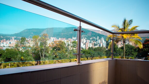 Un balcón con barandilla de vidrio, que ofrece vistas a un paisaje urbano con edificios y palmeras.