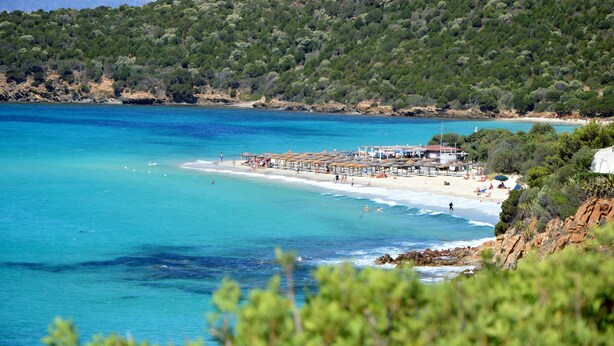 Una playa costera con un muelle, aguas turquesas cristalinas y una ladera boscosa al fondo.