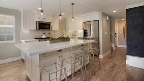 A modern kitchen with a large island, stainless steel appliances, pendant lighting, and a bar stool area. - Indoor/Outdoor Living with Stackable Doors, Firepit, BBQ, Outdoor Heater & More! (Newport Beach)