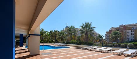 A pool area with lounge chairs, a swimming pool, and a view of palm trees and buildings.