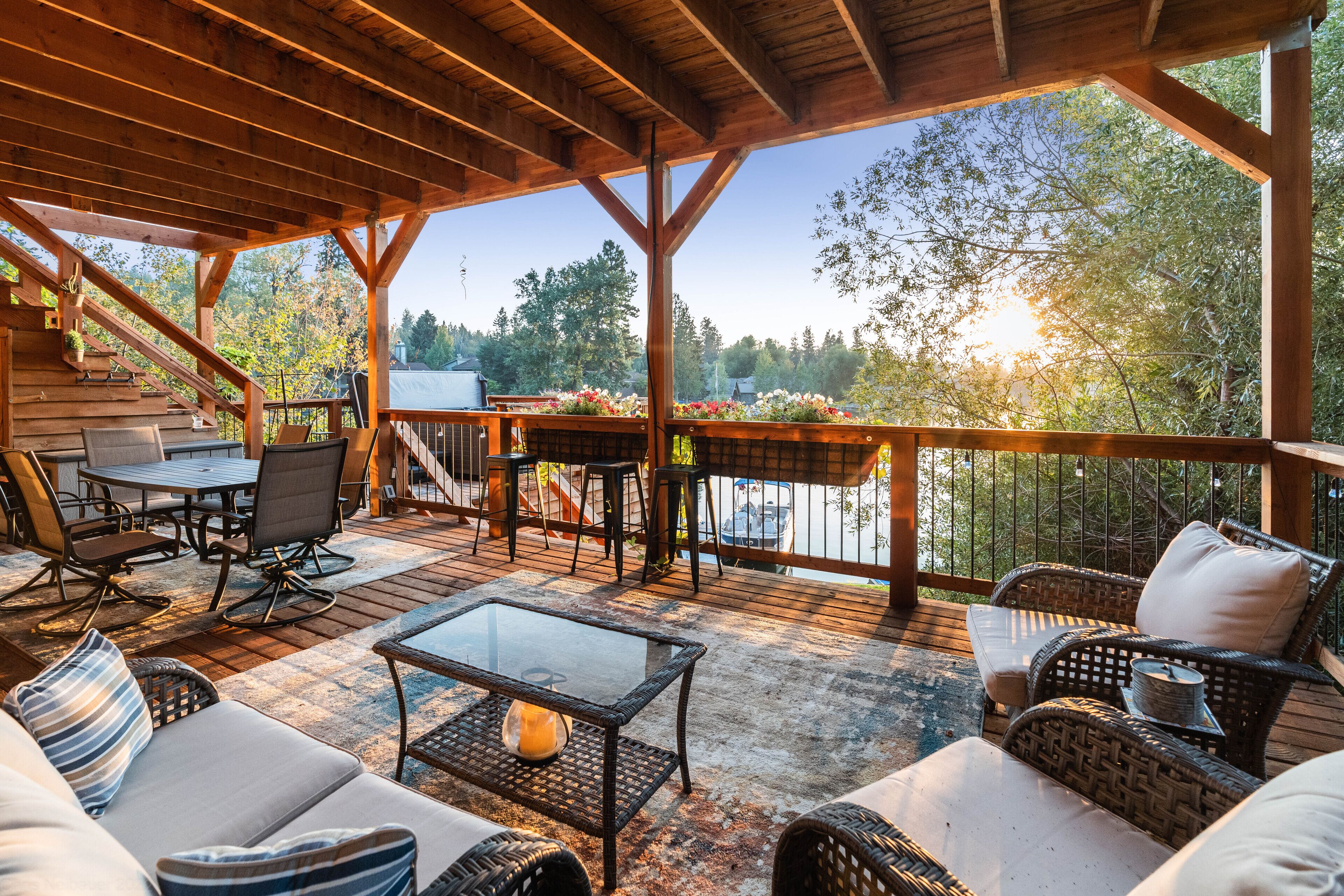 Un patio techado al aire libre con mobiliario de madera, una mesa de vidrio y vistas a árboles y un vehículo al fondo.