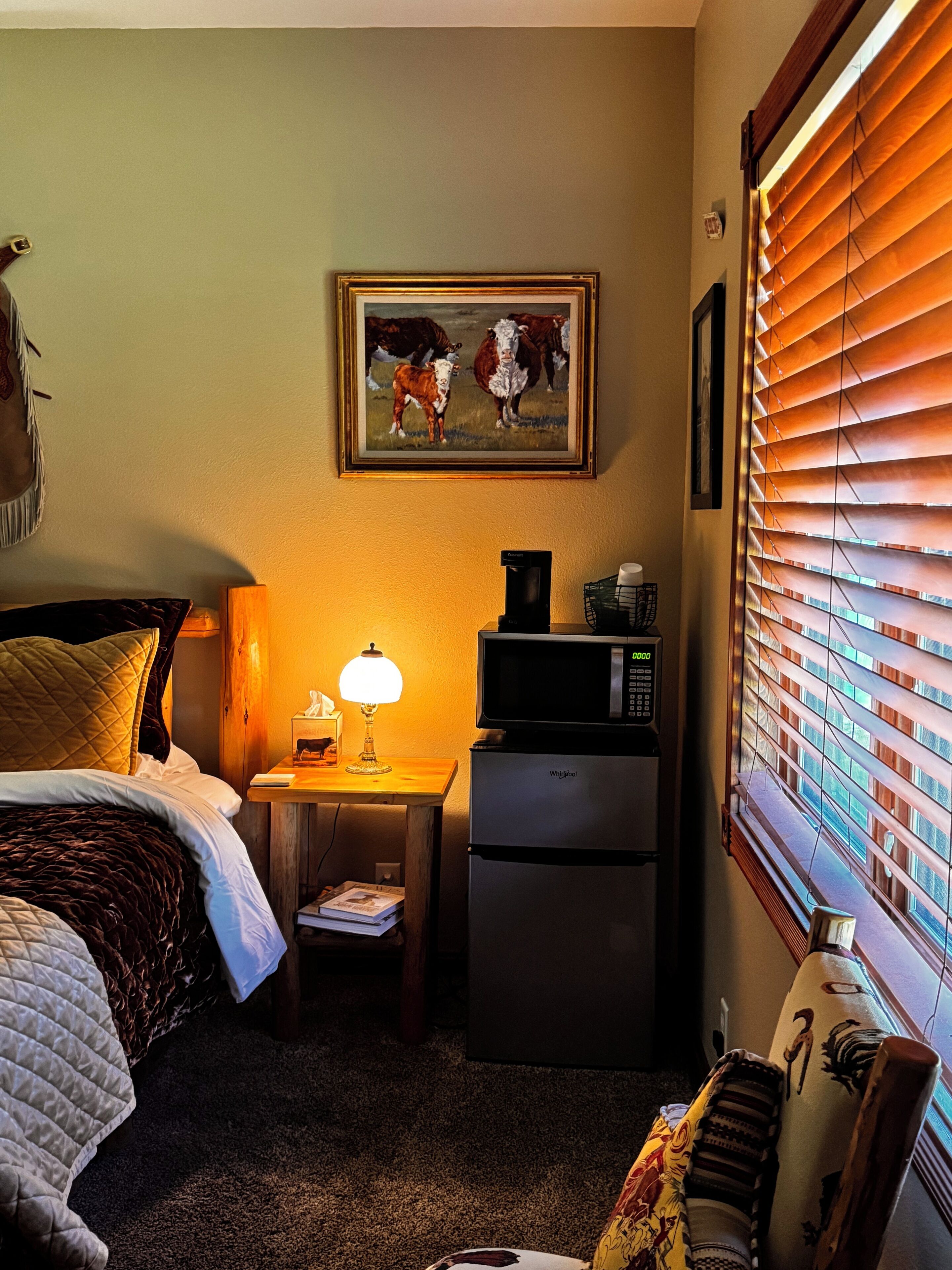 Una camera d'albergo con un letto, una scrivania con una lampada, un frigorifero, un quadro appeso al muro e una finestra con persiane.
