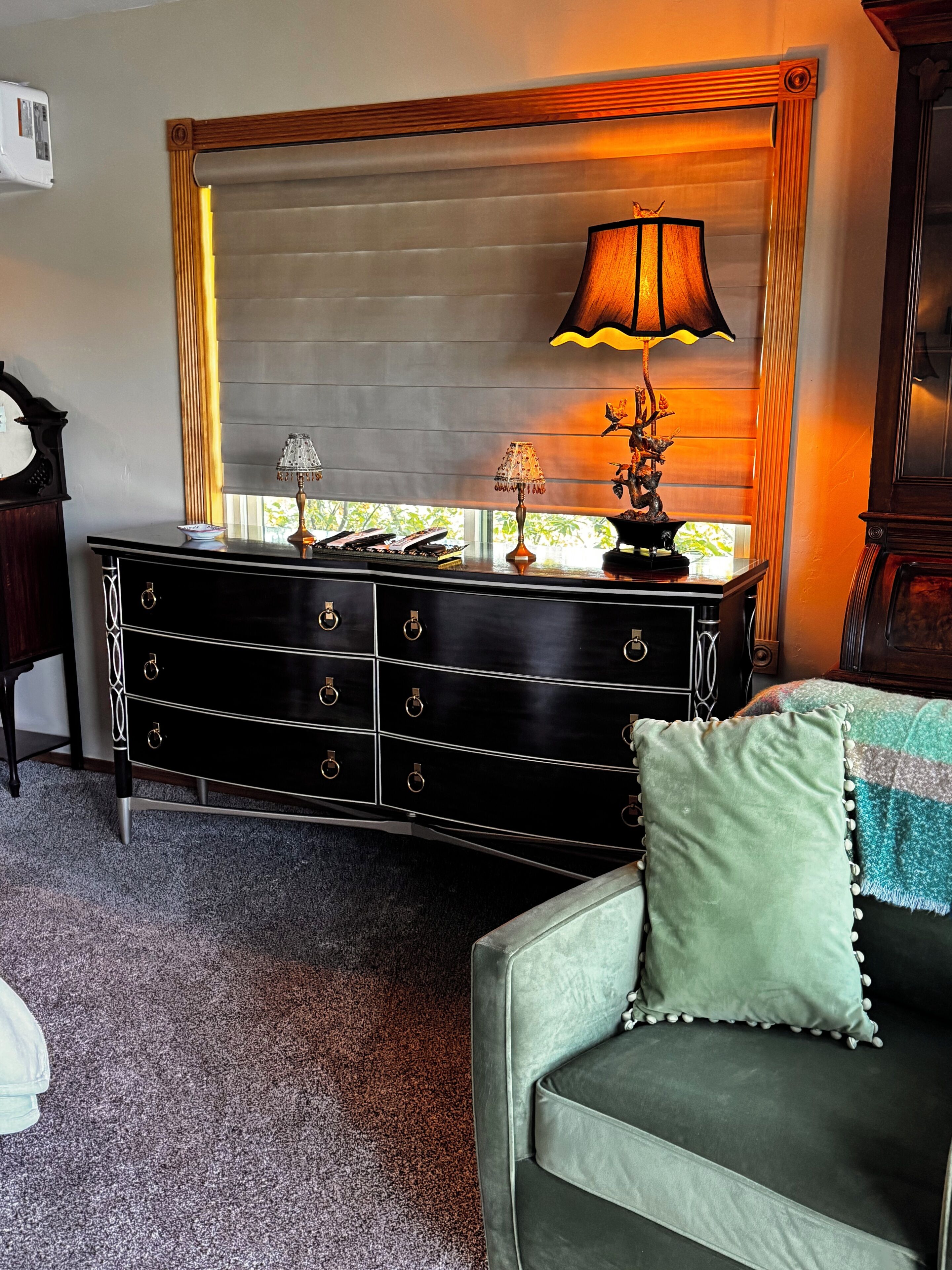 A room with a dark wooden dresser, a green sofa, a lamp, and a window with blinds.