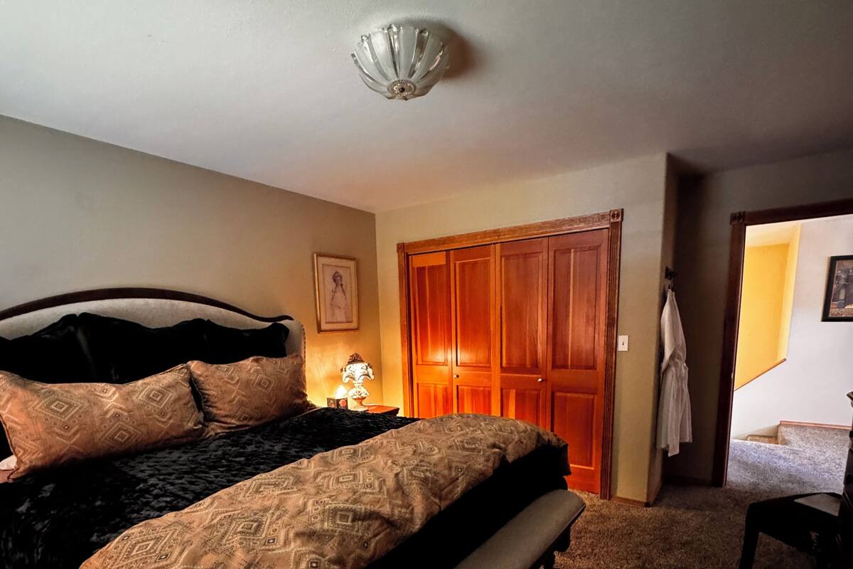 Un dormitorio con una cama, una mesita de noche, una lámpara, una puerta de madera y un pasillo que da a otra habitación.