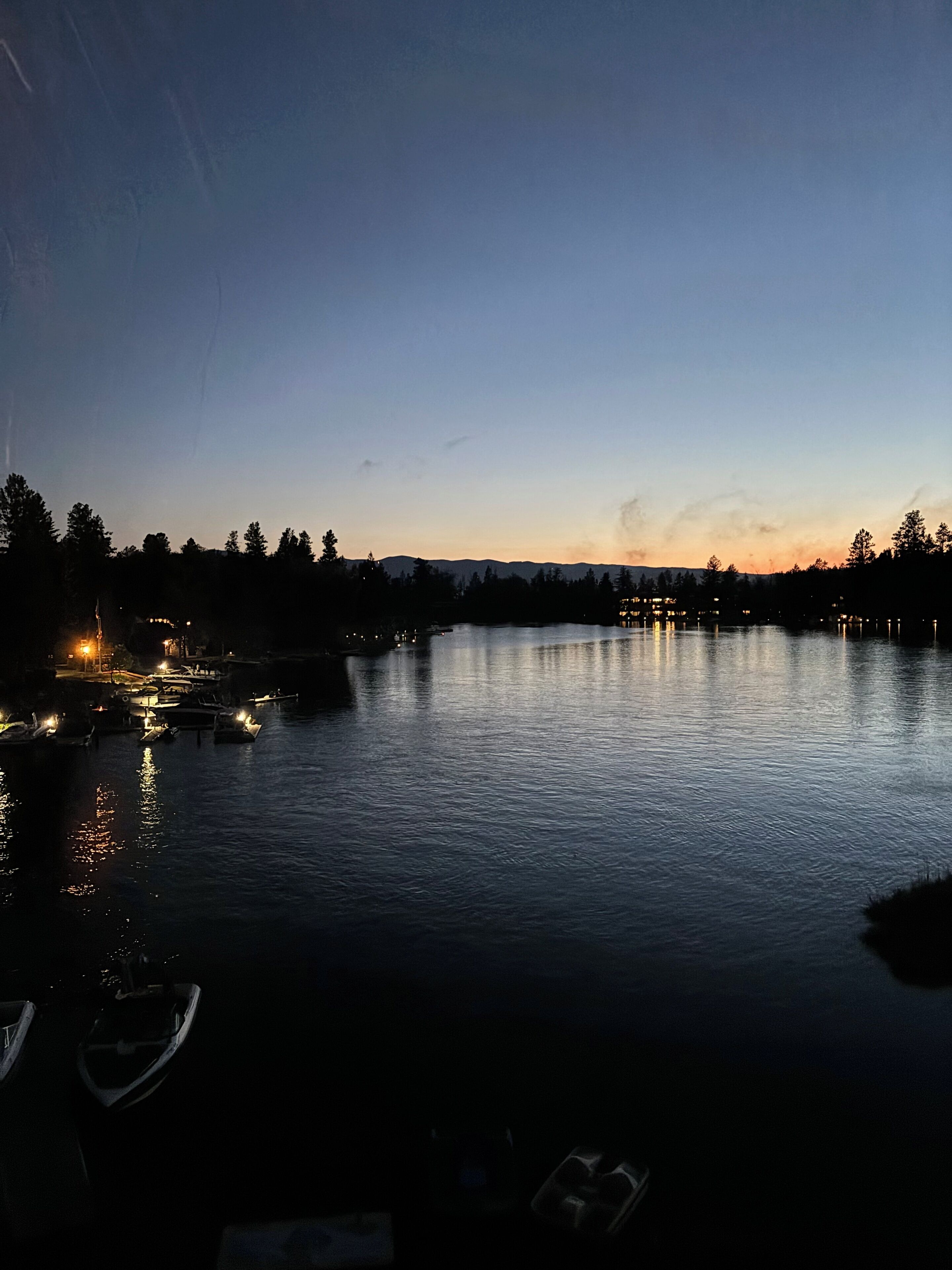 A calm lake at dusk with boats docked on the left and a distant mountain range.
