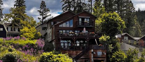 A multi-story wooden cabin with a balcony, situated by a lake with surrounding vegetation.