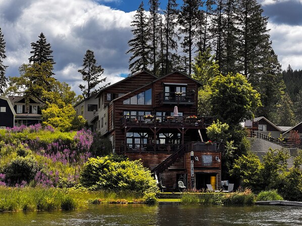 Une cabane en bois à plusieurs étages, dotée d’un balcon, située au bord d’un lac, entourée de végétation.