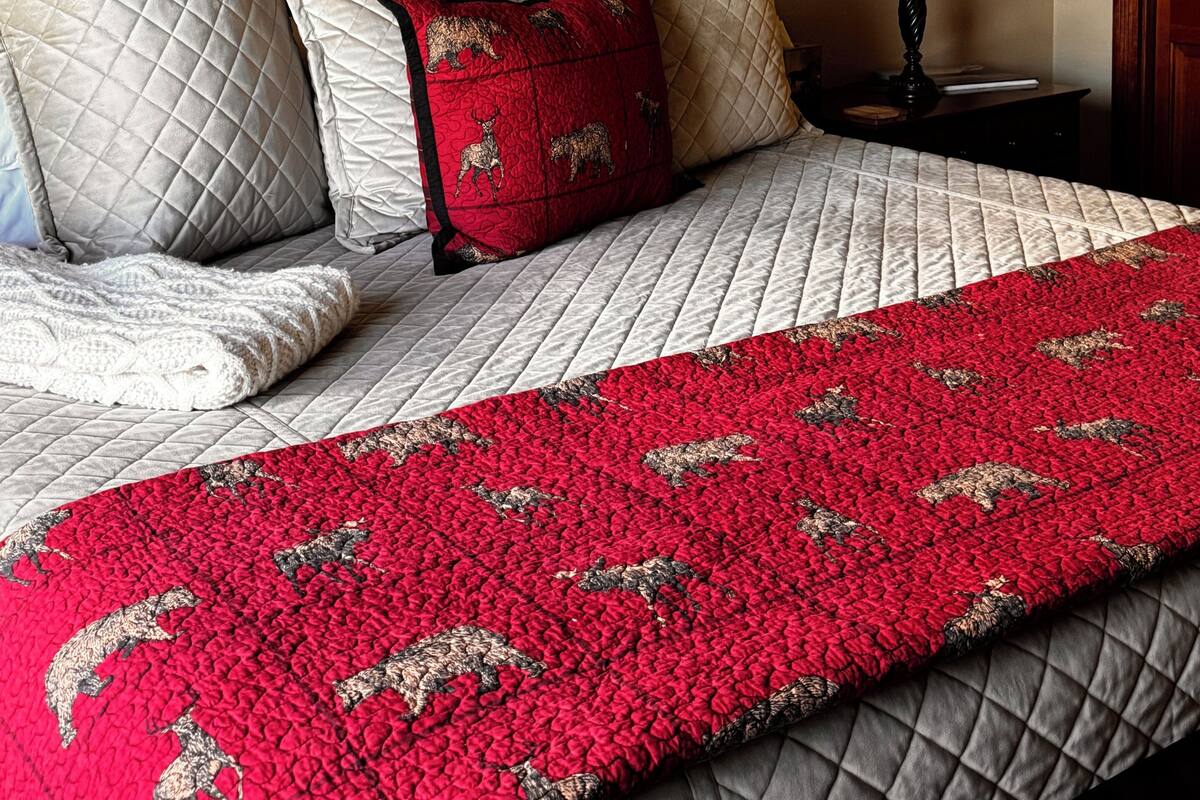 Una cama con colcha, almohadas, una manta roja, un estante en la pared con adornos, un cuadro enmarcado y una lámpara de noche.