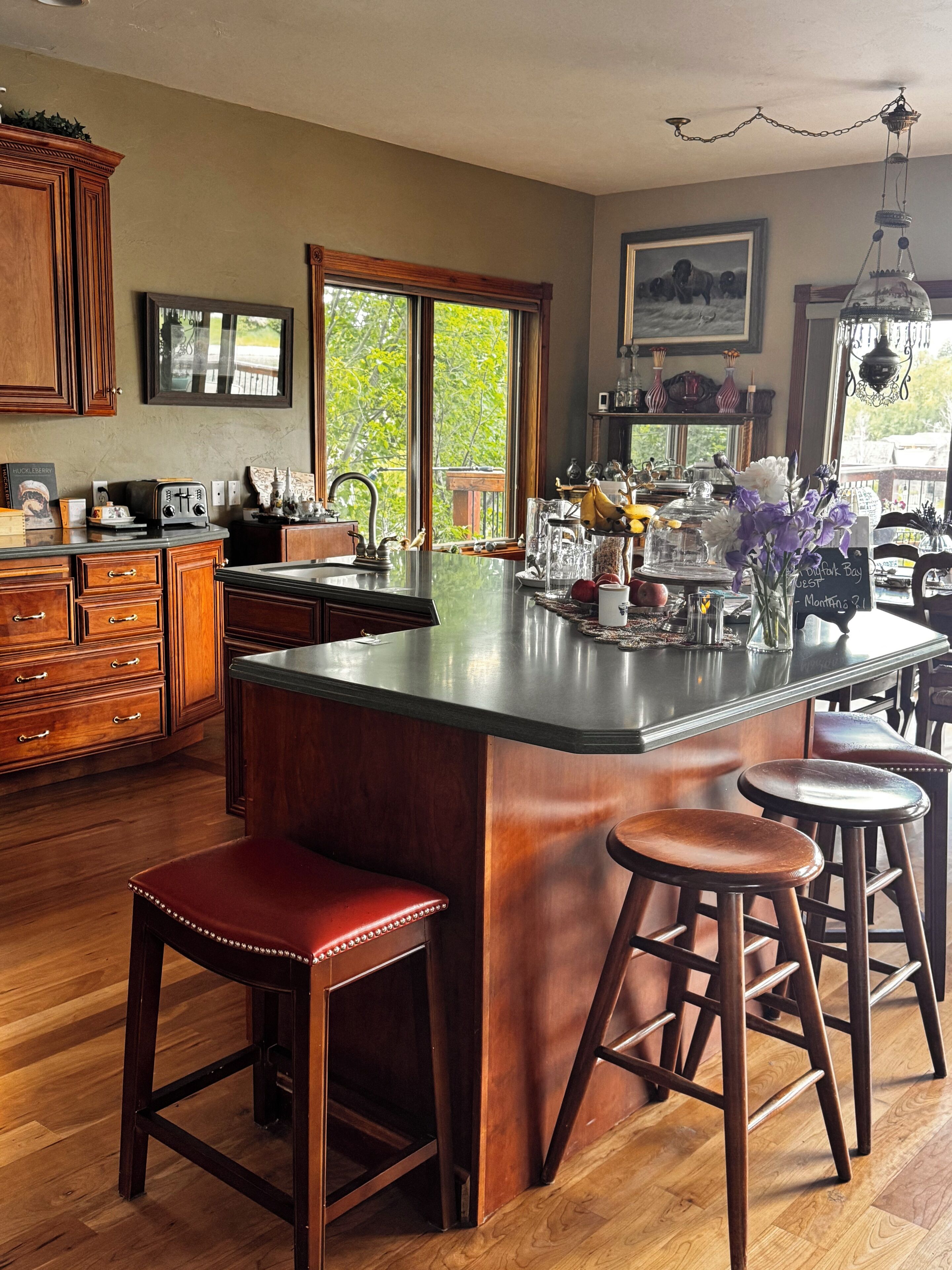Una cocina con armarios de madera, una isla grande con taburetes, un ventanal con vistas y diversos utensilios de cocina.