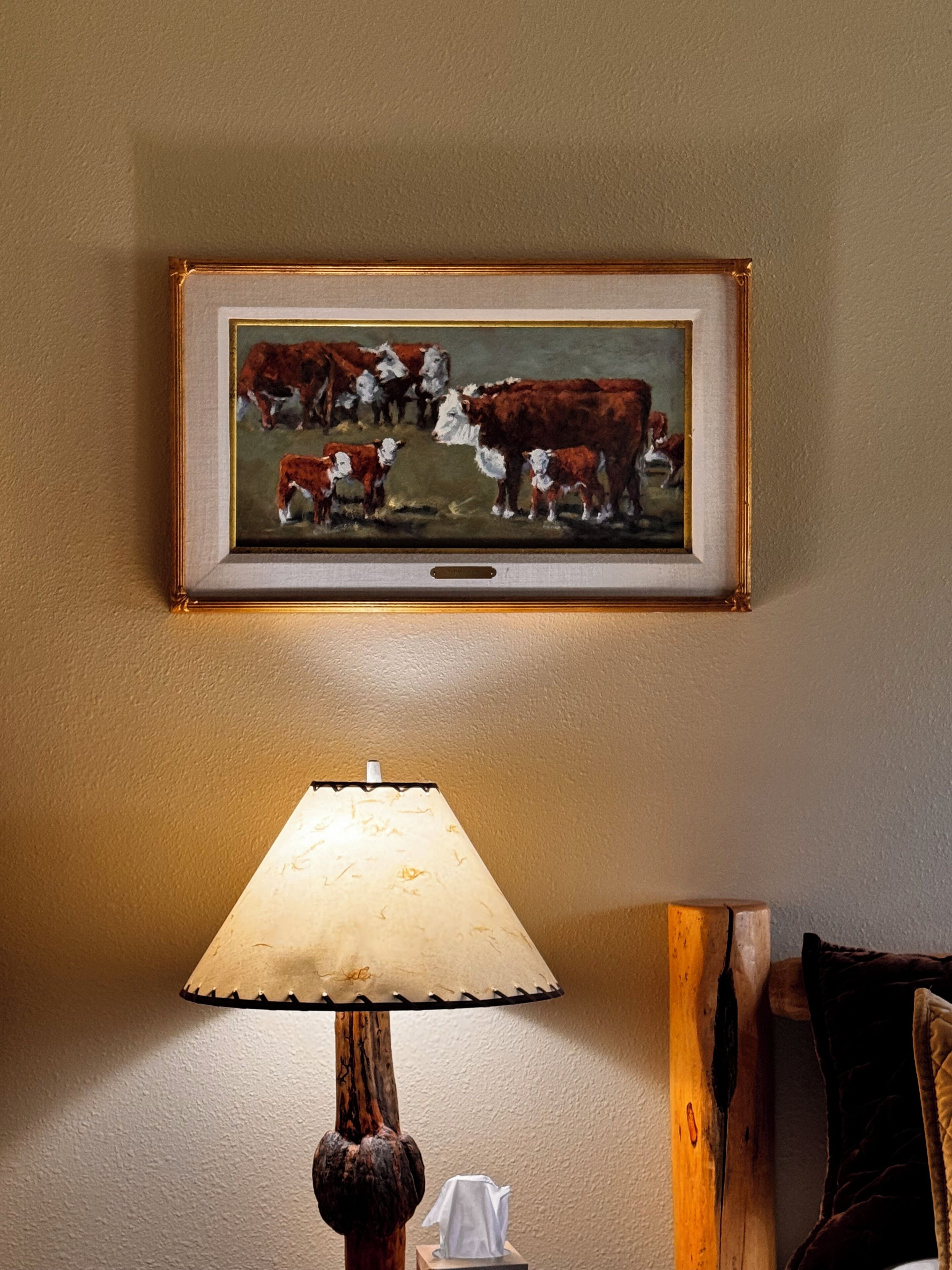 Um quadro com vacas pendurado na parede, uma luminária de madeira e uma mesa de cabeceira com um estojo de lenços.
