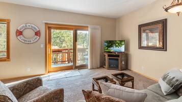 Ein Wohnzimmer mit einer Couch, einem Couchtisch, einem Fernseher und einem Kamin. Eine Glastür führt auf eine Terrassenanlage im Freien.