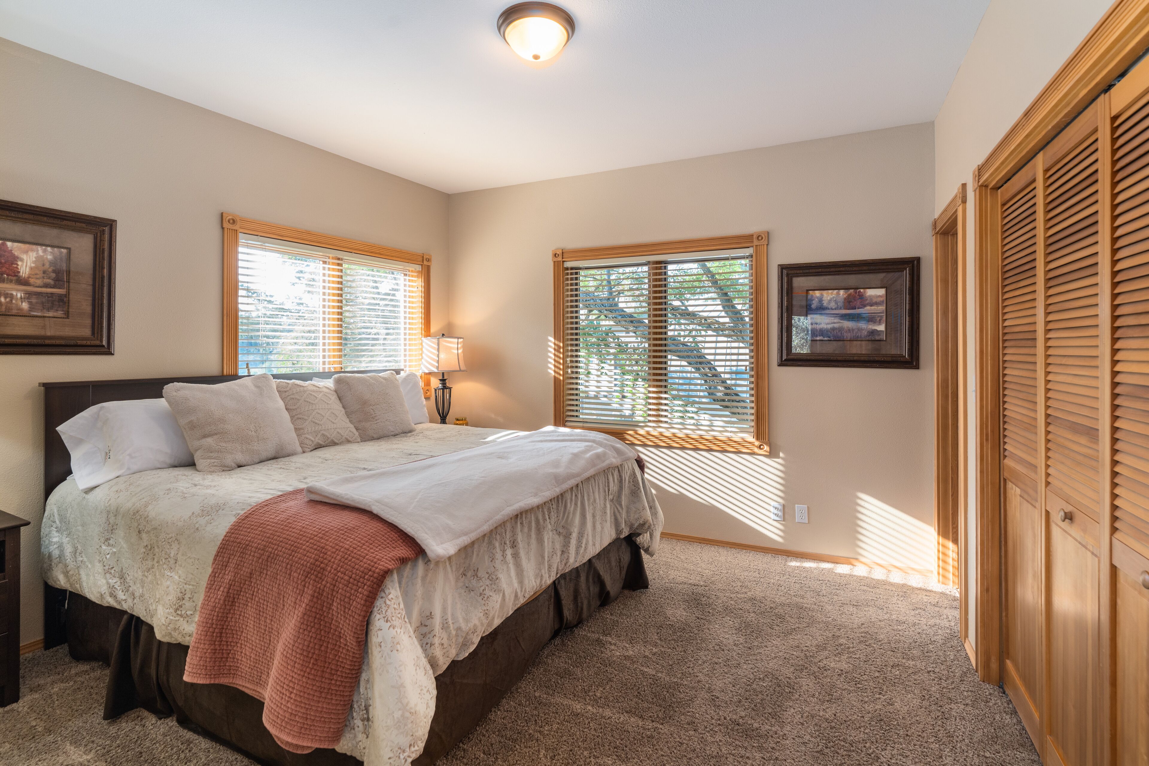 A bedroom with a large bed, two windows with blinds, a nightstand, a lamp, and a wardrobe.