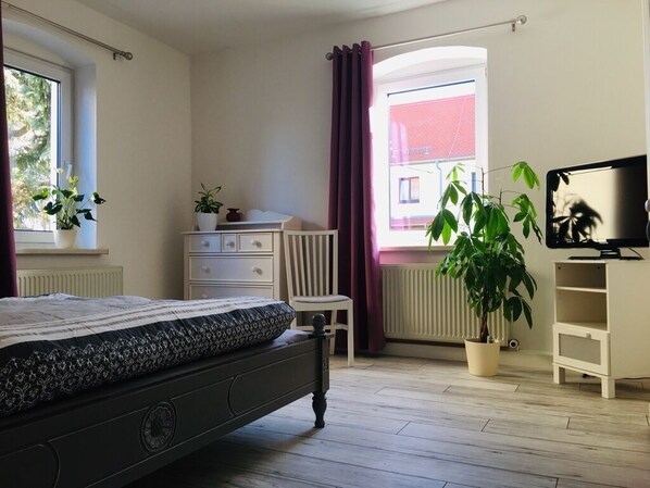 Un dormitorio con cama, cómoda, silla, televisión y ventana con cortinas.