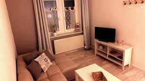 A living room with a sofa, a TV on a stand, a window with curtains, and a radiator. - Living in a country house near Bautzen/ OT Doberschau (up to 4 persons; vacation apartment) (Doberschau)