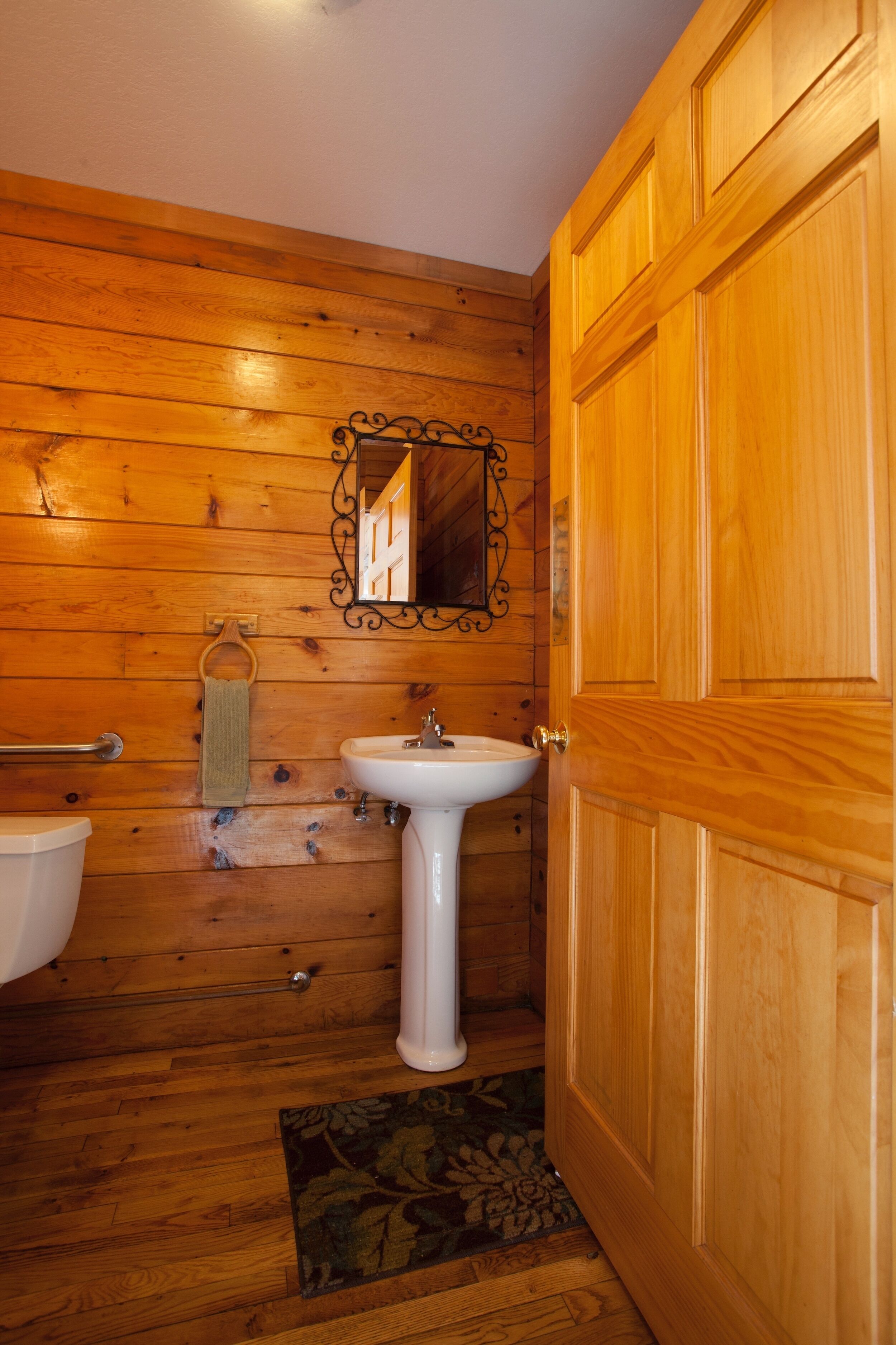 Un baño con paneles de madera, un lavamanos blanco de pedestal, un perchero y una puerta de madera.