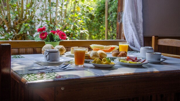A breakfast table with a variety of food items, a vase of flowers, and a view of greenery through the window. - Pousada Cruzeiro do Sul (Itatiaia)