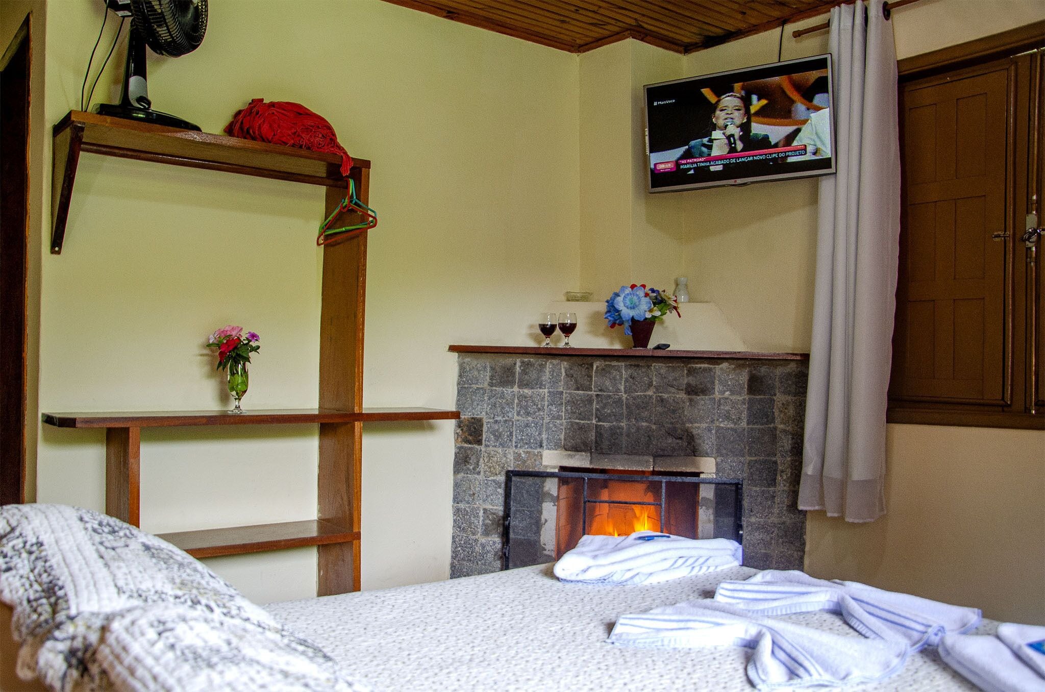 Una habitación con una cama, una chimenea de piedra, un televisor montado en la pared y un estante de madera con un objeto rojo.
