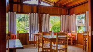 A wooden dining area with a table and chairs, large windows with curtains, and a view of greenery outside. - Pousada Cruzeiro do Sul (Itatiaia)