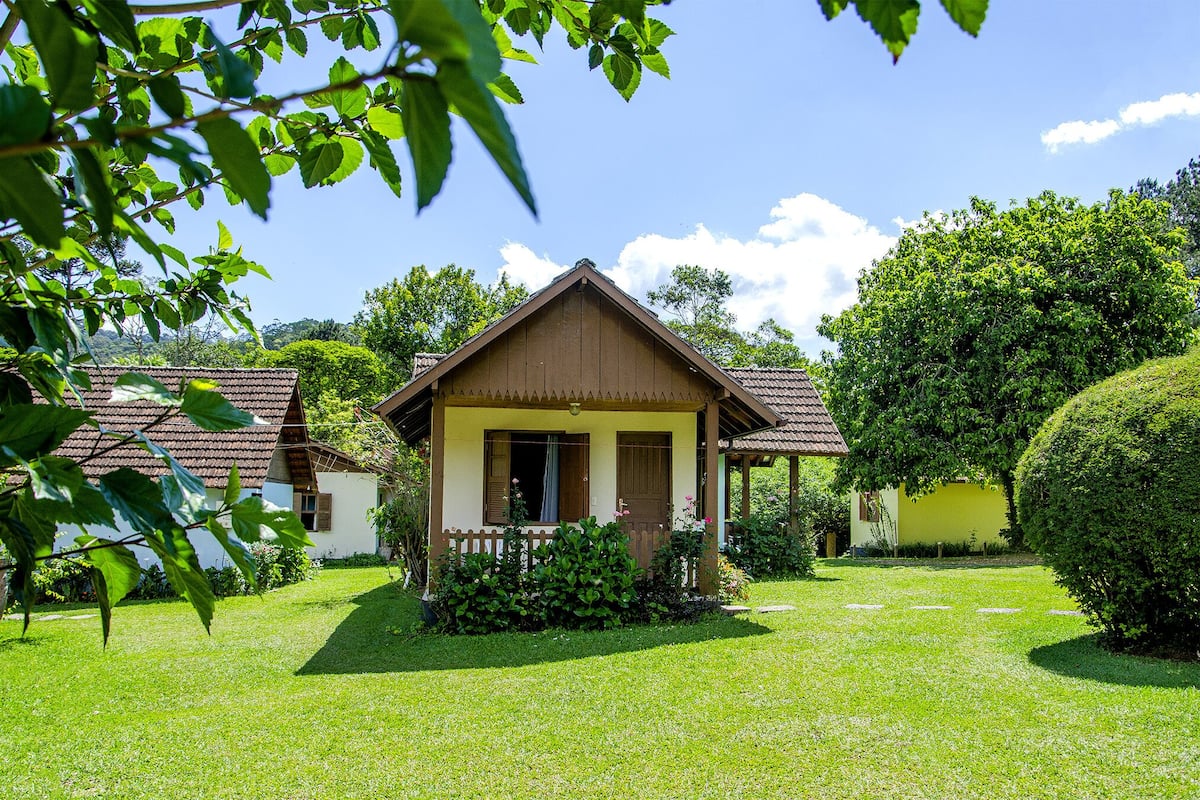 Une petite maison de plain-pied, avec un toit brun et des murs blancs, entourée d’une pelouse bien entretenue et de divers arbres.