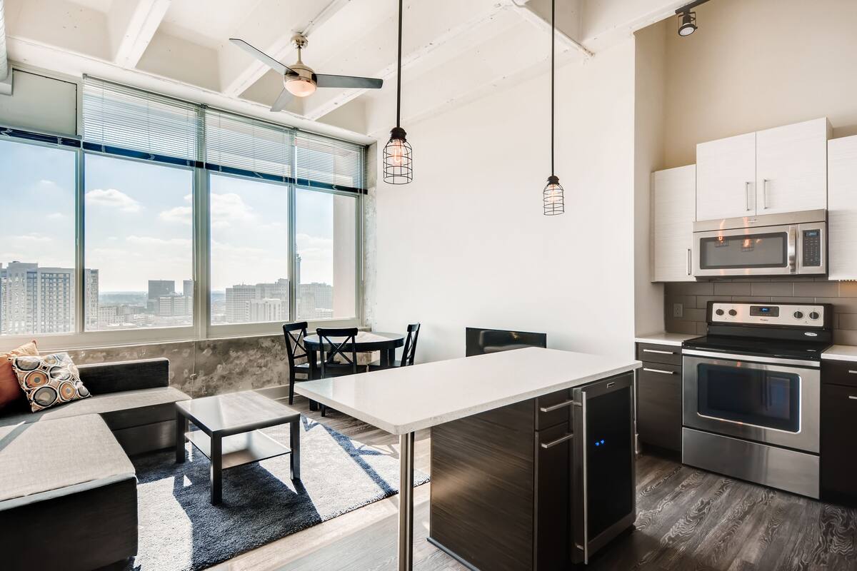 A modern kitchen with stainless steel appliances, a dining area with a white table and chairs, and a large window offering a city view.