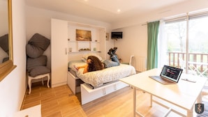 Een slaapkamer met een bed, een bureau met een laptop, een stoel, een televisie en een raam met gordijnen.