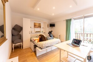 Une chambre à coucher avec un lit, un bureau avec un ordinateur portable, une chaise, une télévision et une fenêtre avec des rideaux.