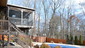Une terrasse avec un escalier menant à une zone abritée, un trampoline et une cour arrière clôturée avec des arbres.