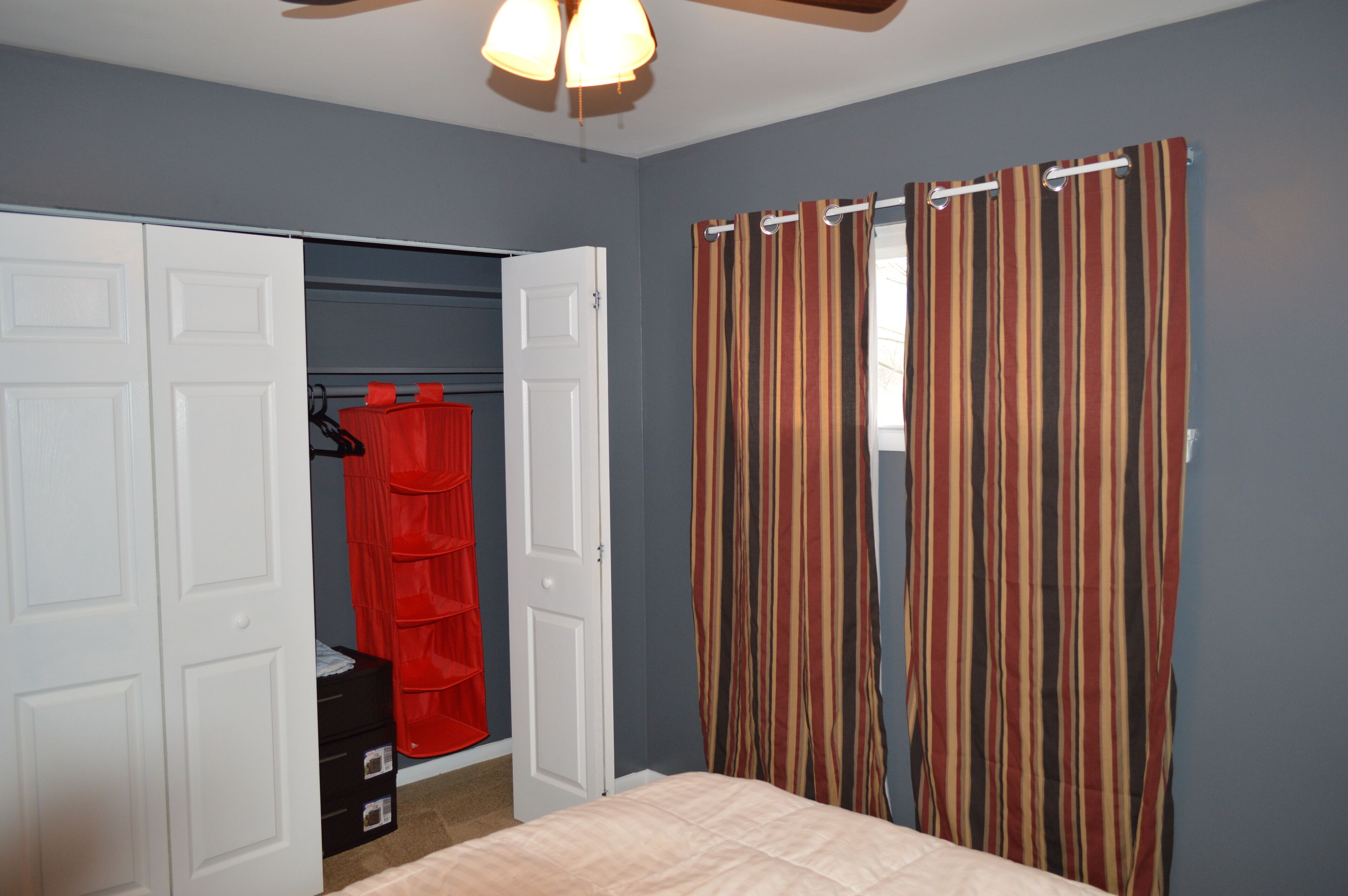 A bedroom with a bed, a closet with a red hanging organizer, and a striped curtain.