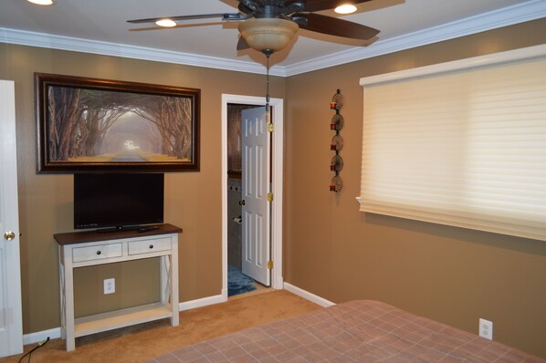 A bedroom with a ceiling fan, a television on a white console table, a door leading to a bathroom, and a window with blinds. - Comfy, Serene Home on Quiet Cul de Sac, Private backyard pool
Lic # STR23-00110 (Rockville)