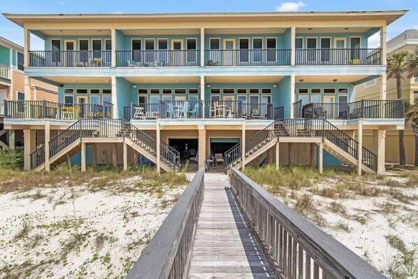 A multi-story beachfront hotel with a central entrance, balconies, and a wooden boardwalk leading to the entrance.