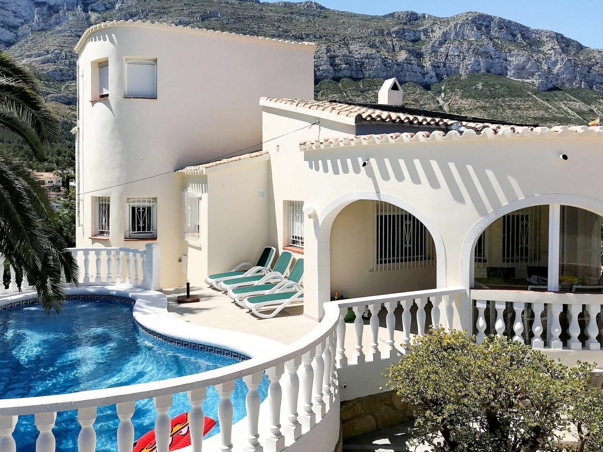 A white villa with a swimming pool, a balcony with lounge chairs, and a view of rocky mountains.