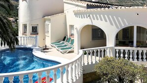 A white villa with a swimming pool, a balcony with lounge chairs, and a view of rocky mountains.