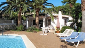 Un espace piscine doté de chaises longues et d’une petite table de patio, entouré de palmiers et d’un bâtiment blanc avec un toit en tuiles.