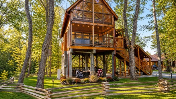 A multi-level wooden cabin with a balcony, surrounded by trees and a well-maintained lawn.