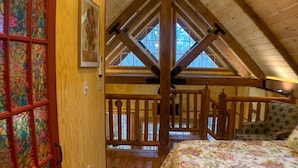 A bedroom with a wooden ceiling, a bed with a floral bedspread, a wooden door with stained glass, and a view of the outdoors through a window.