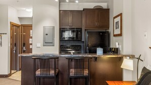 A modern kitchen with dark wood cabinets, a built-in microwave, and a coffee machine. There are two bar stools and a small wooden table. - Time for a Getaway? Pool and A/C (Fernie)