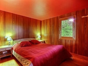 Quarto com painéis de madeira, uma cama, duas mesas de cabeceira e uma janela com vista para o verde.